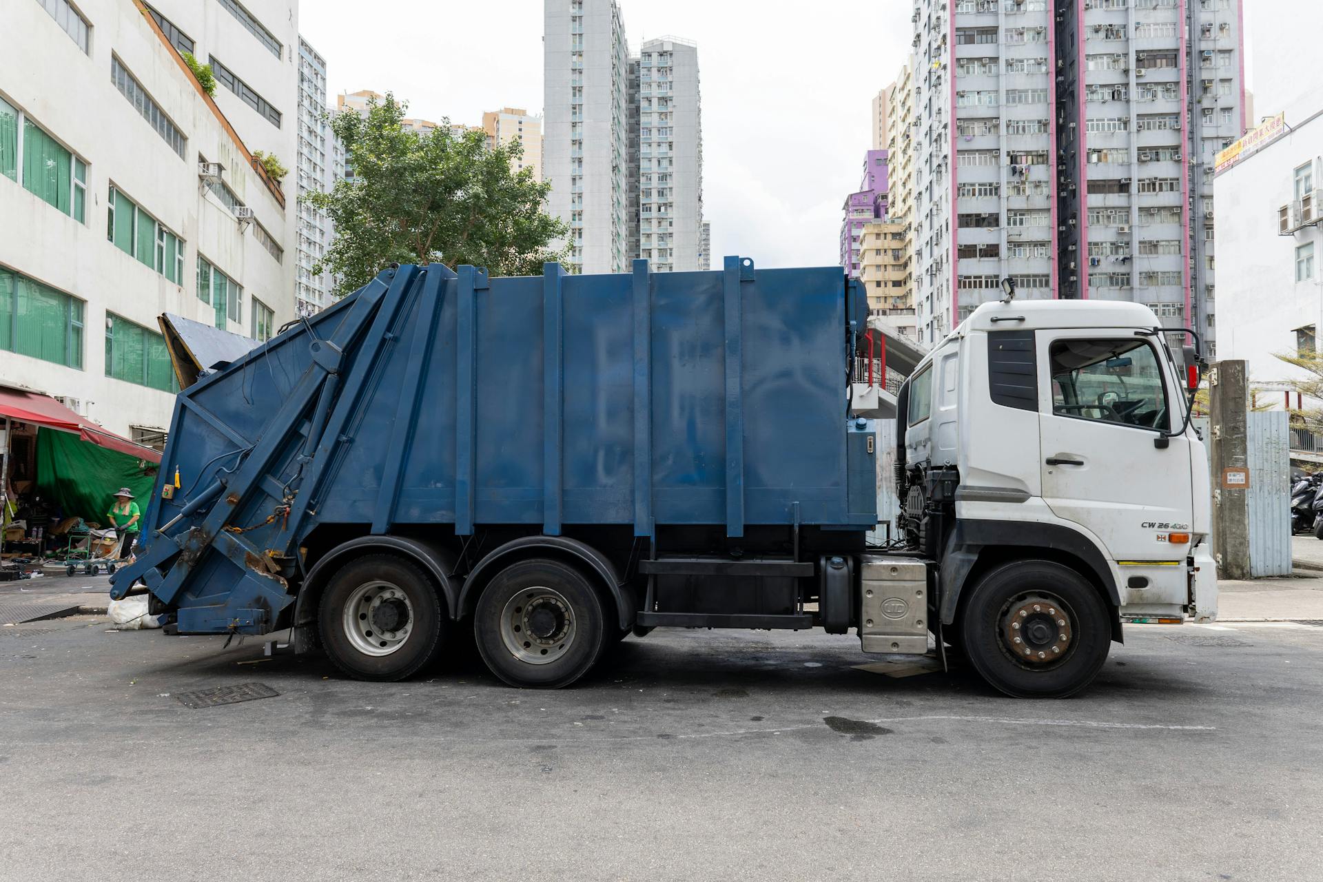 A blue garbage truck parked in a vibrant city street surrounded by tall buildings.
