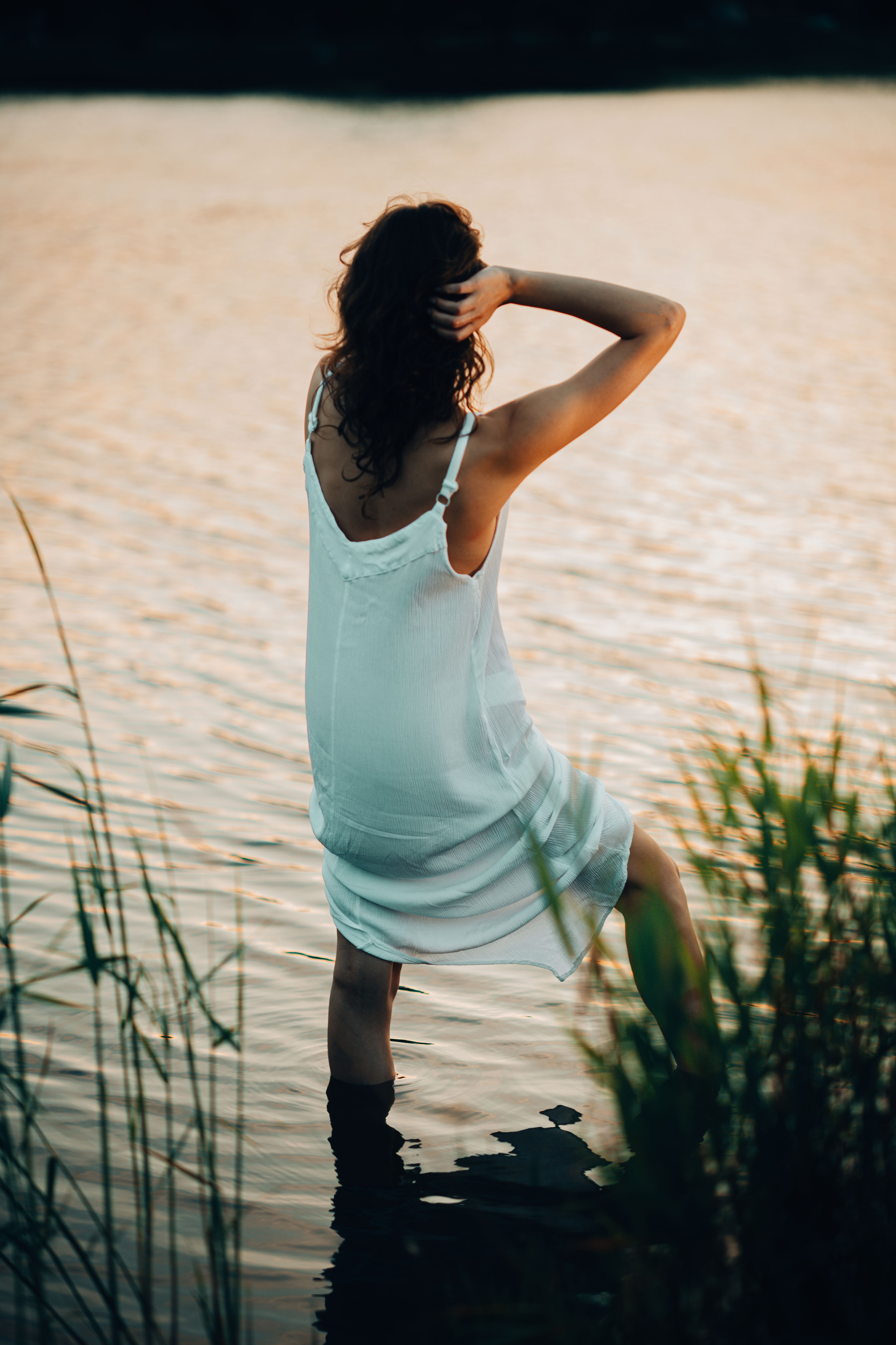 A woman in a white dress standing in a calm lake enjoying a serene sunset moment.