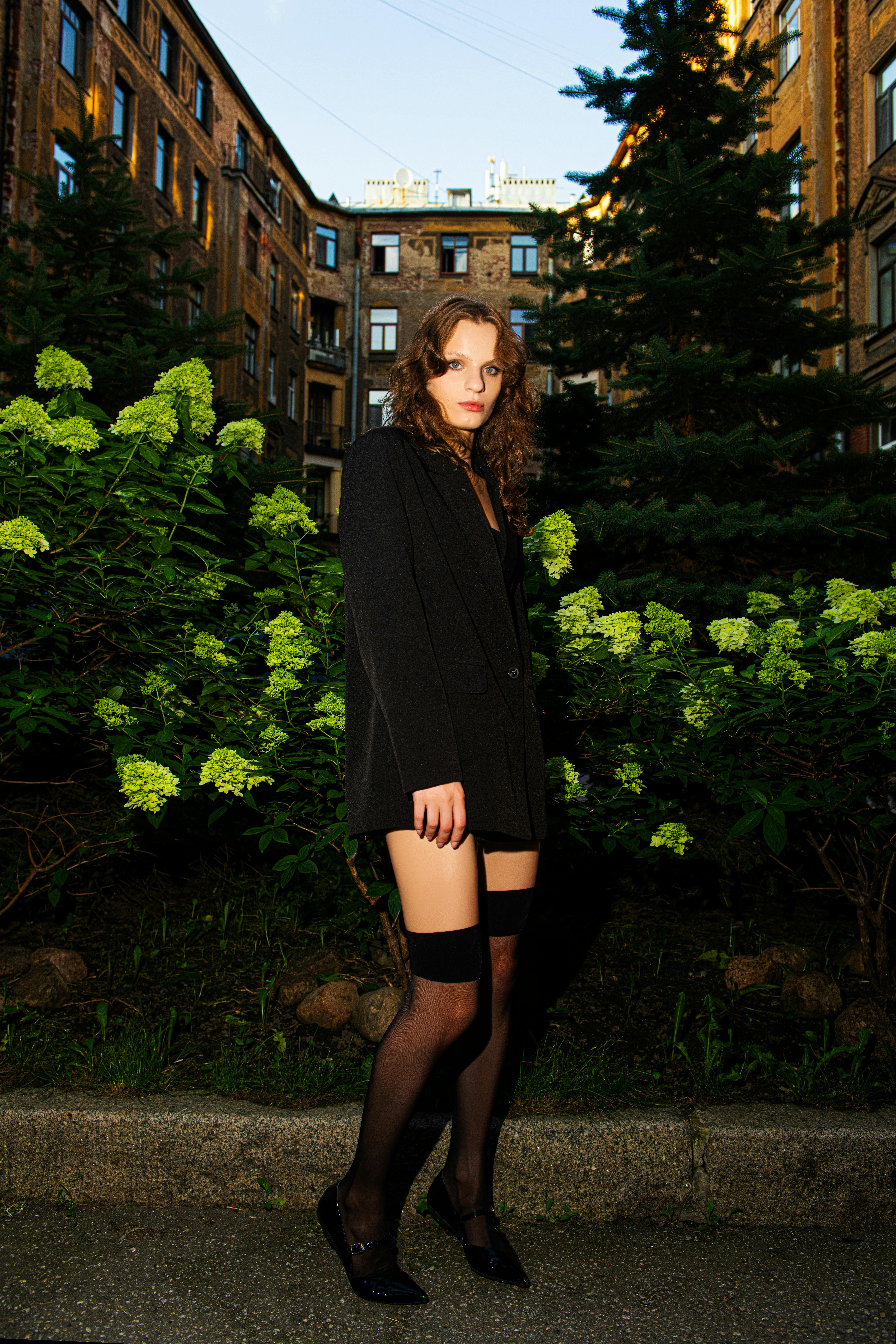 Chic woman in black attire posing in an urban garden at dusk with lush greenery and historic buildings.