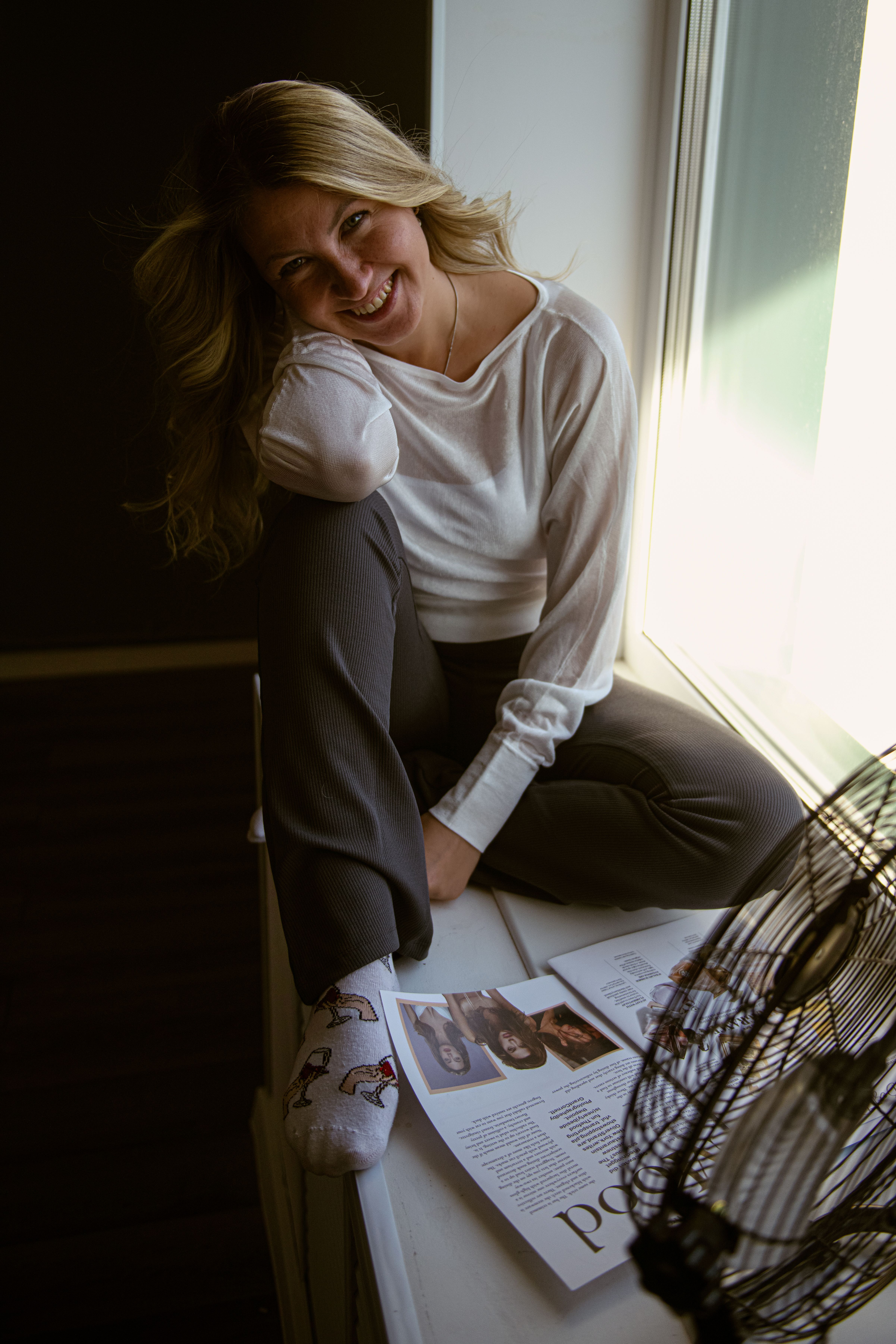 Woman in relaxed pose by a window, smiling with a magazine and a fan.