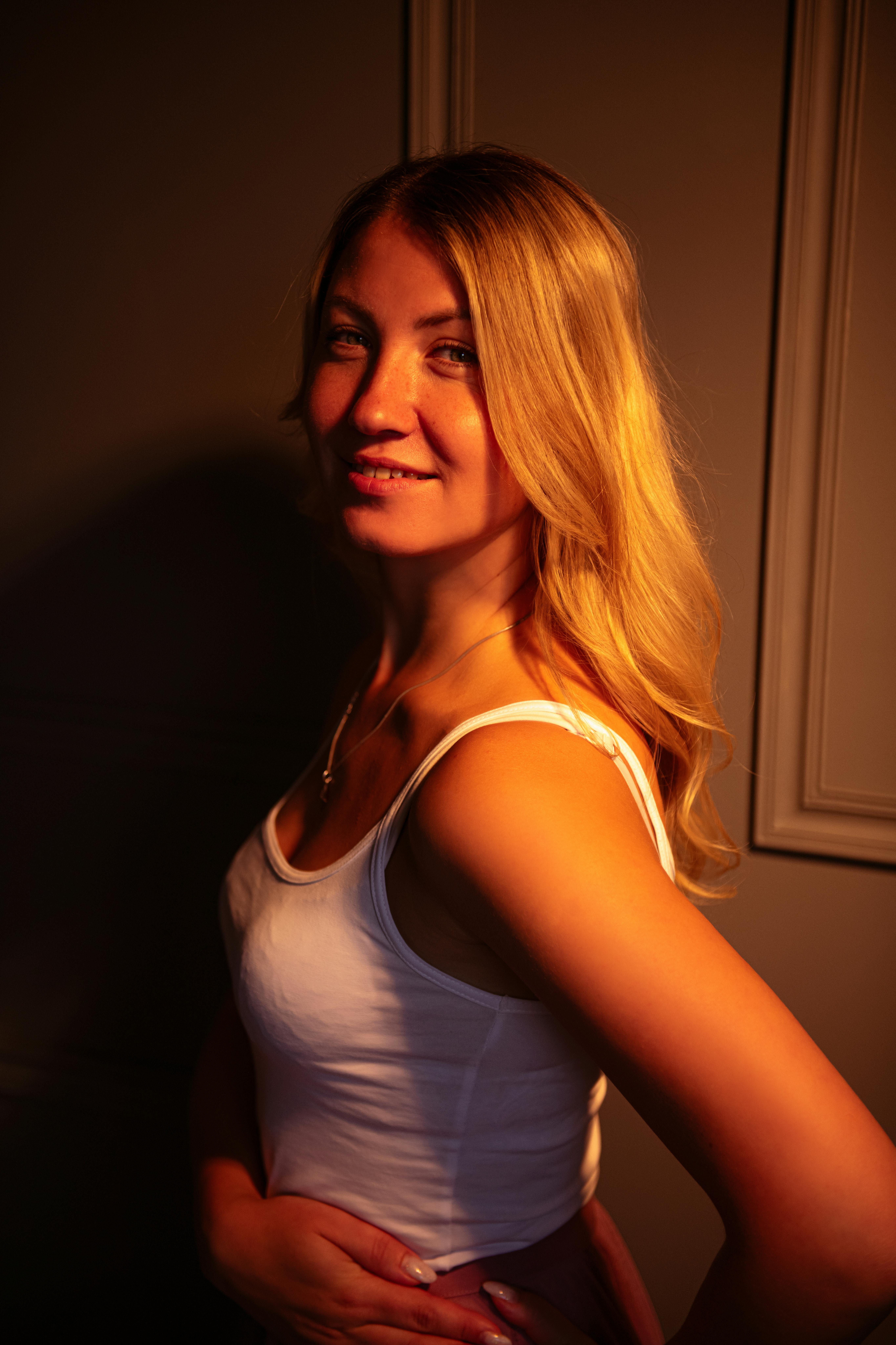 A blonde woman in a white top smiles warmly standing indoors with gentle lighting.