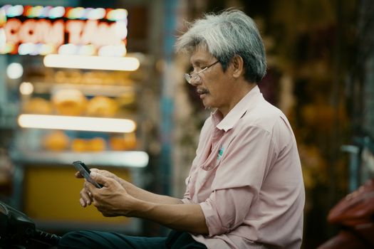 Senior man sitting outdoors using smartphone with blurred city lights in background.