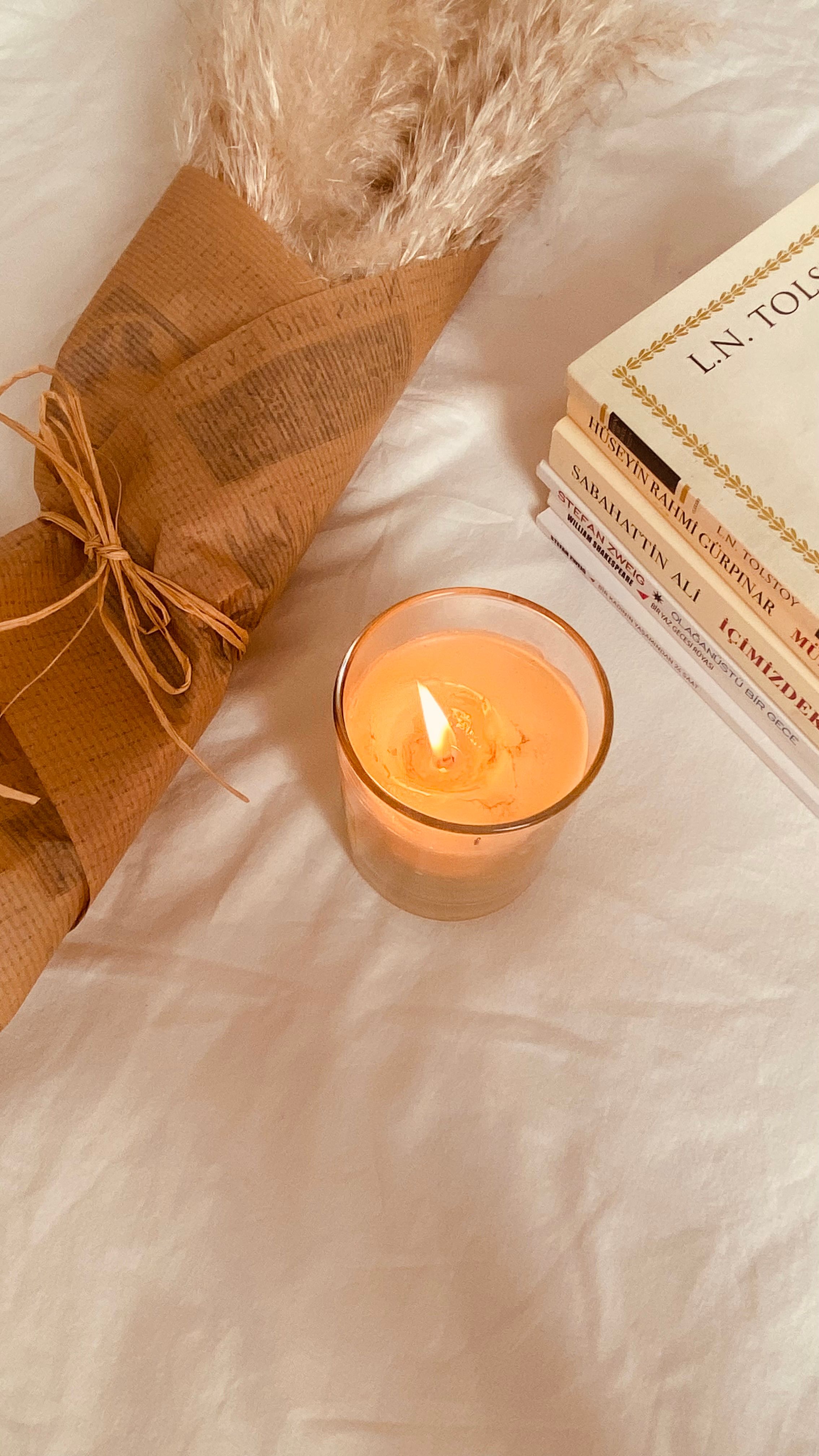 Warm ambiance with a lit candle, books, and pampas grass on a bed.