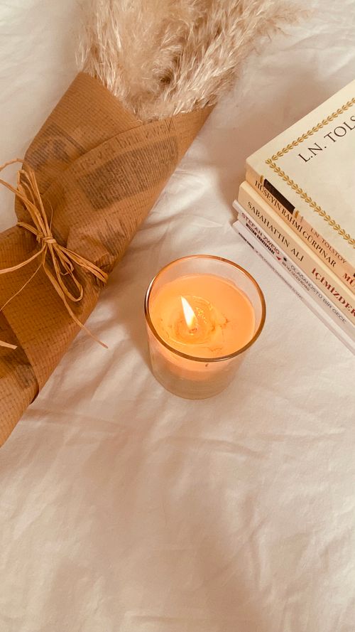 Free Warm ambiance with a lit candle, books, and pampas grass on a bed. Stock Photo