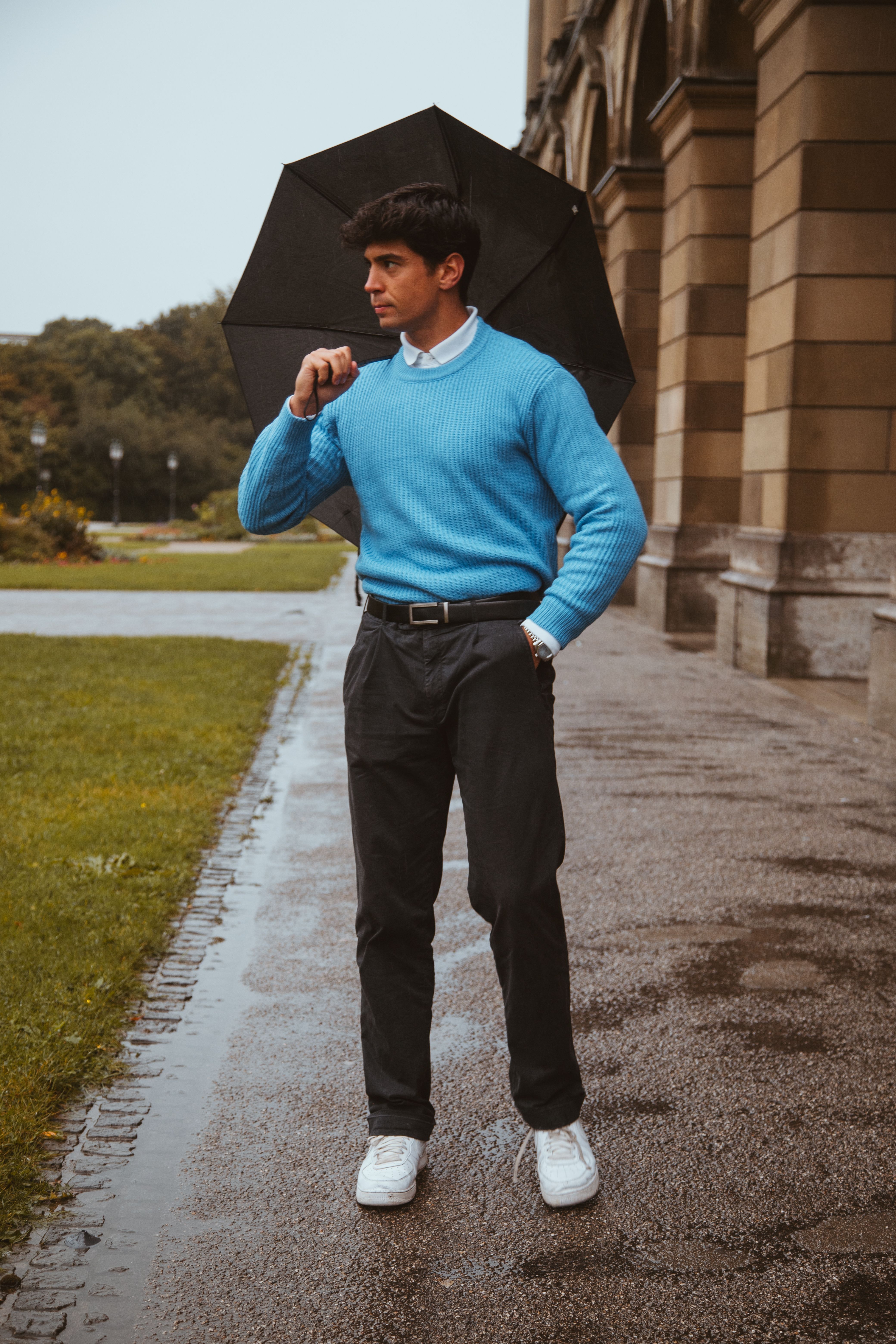 A fashionable man in a blue sweater walks with an umbrella on a rainy day in Munich.