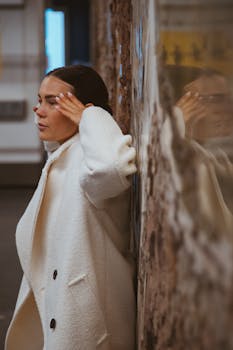 A woman in a white coat leans against a wall indoors, reflecting deeply.