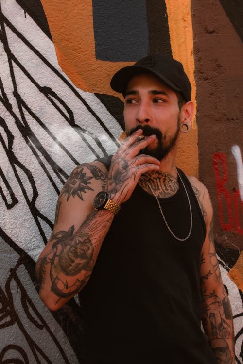 Free Street style portrait of a tattooed man smoking, standing against a graffiti-covered wall. Stock Photo