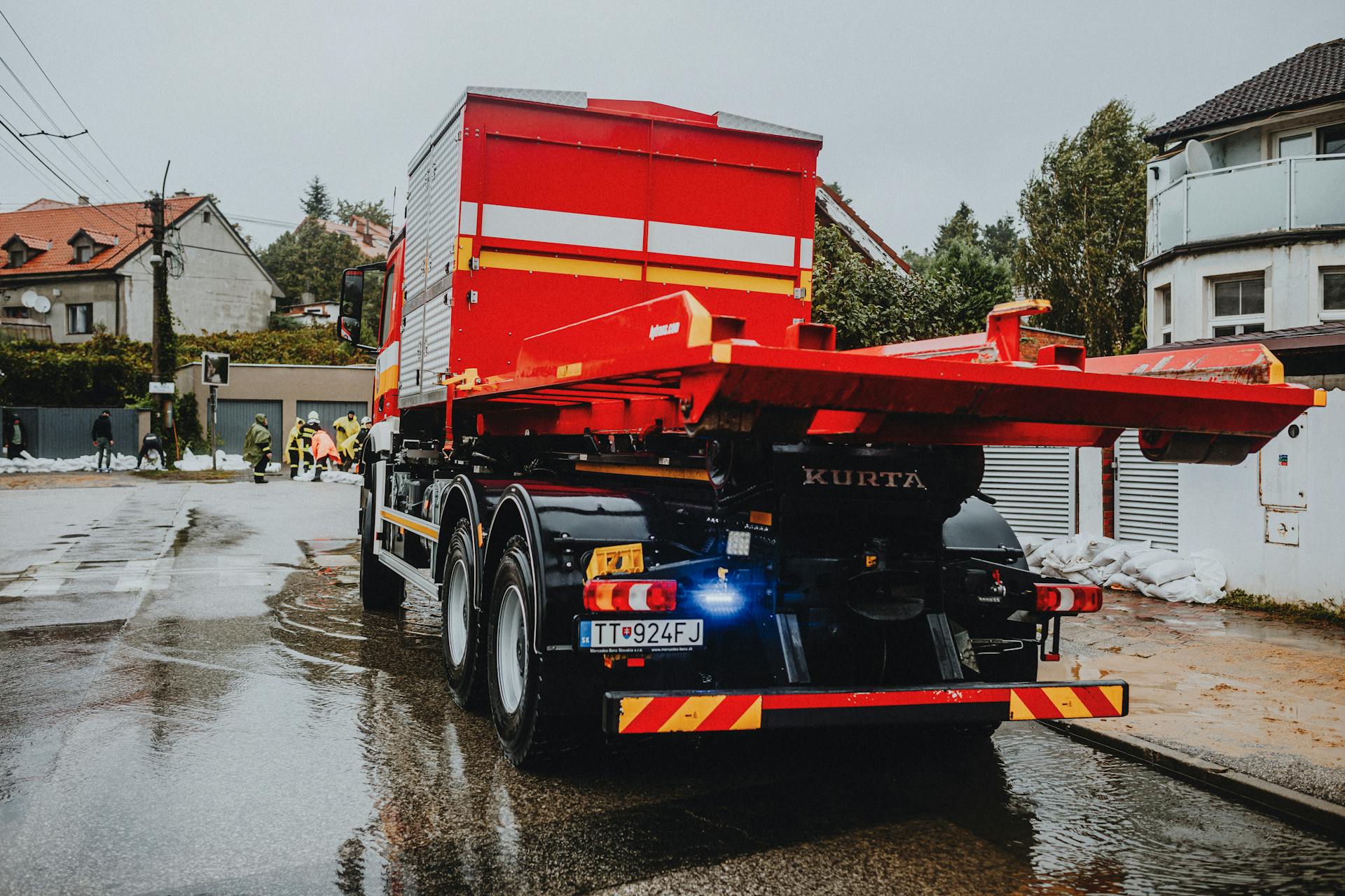 A red tow truck parked on a wet street in an urban neighborhood with workers in the background.