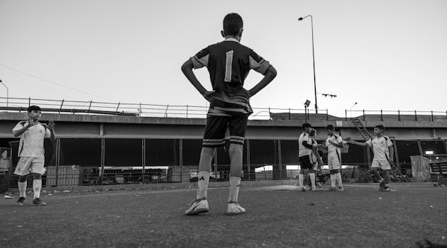 Black and white photo of boys playing soccer in an urban setting under a bridge.
