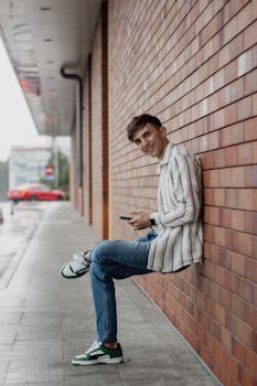 Casual young man with a smile sitting against a brick wall, using phone.