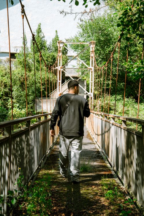 Free A man walks across a rustic suspension bridge surrounded by greenery during the day. Stock Photo