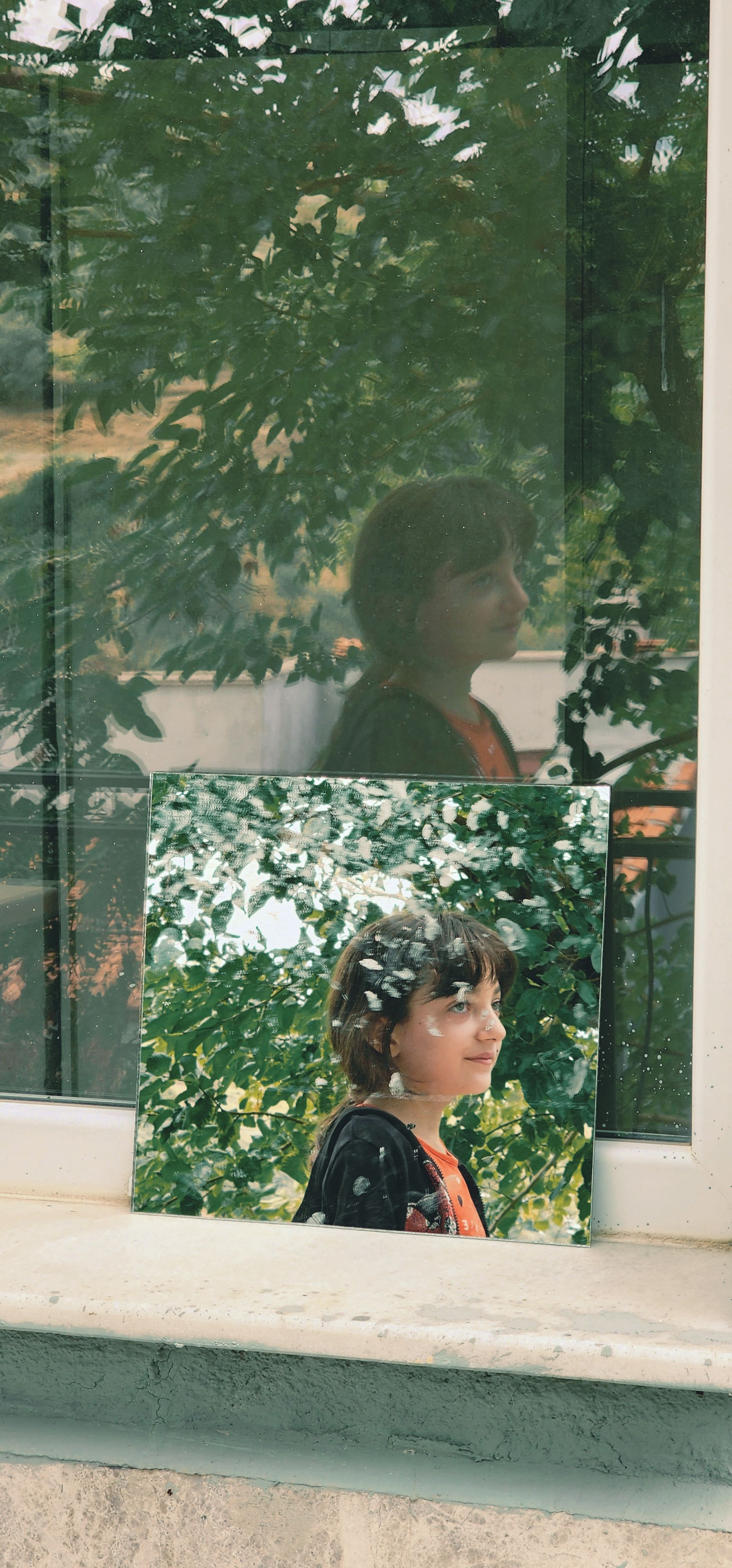 A thoughtful child reflected in a window next to their portrait, outdoors.