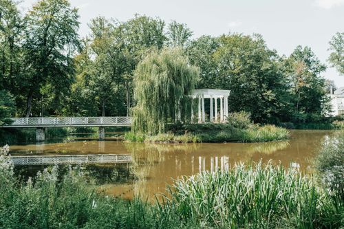 Free Scenic view of a white gazebo by a pond surrounded by lush greenery in a peaceful park setting. Stock Photo