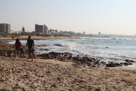 A scenic view of people walking along Gqeberha Beach with city skyline in the background.