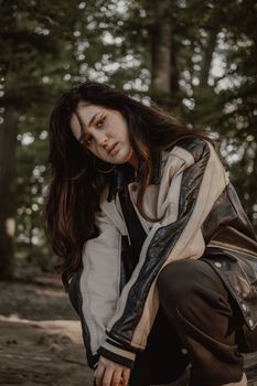 A woman poses in a forest wearing a leather jacket, surrounded by trees