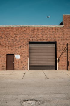 A brick industrial building with a garage door and airplane in the clear sky above.