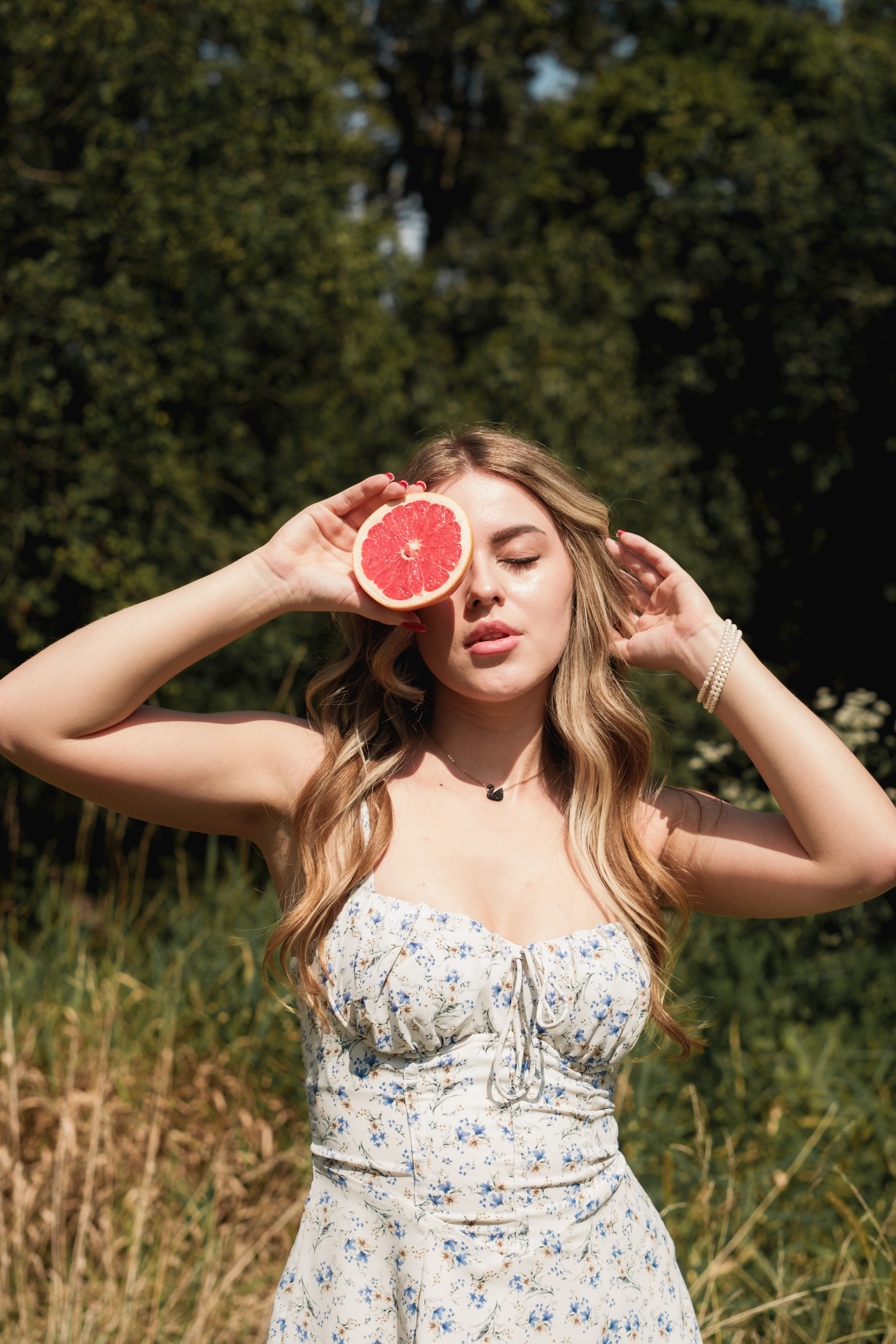 A young woman in floral dress holds a grapefruit slice outdoors in summer.