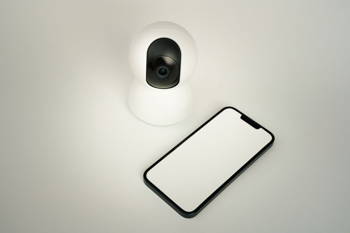 Câmera inteligente branca posicionada ao lado de um smartphone sobre uma superfície clara.