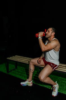 Muscular man takes a break during workout, seated on bench, drinking from a shaker in a dark gym setting.