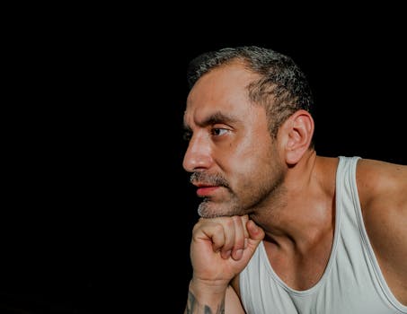 Close-up portrait of a man in a white tank top with a thoughtful expression against a dark background.