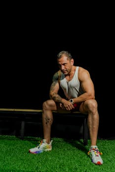 A muscular man in athletic wear rests on a bench in a gym with black background.