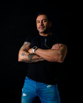 Tattooed man posing confidently with crossed arms against a dark background.