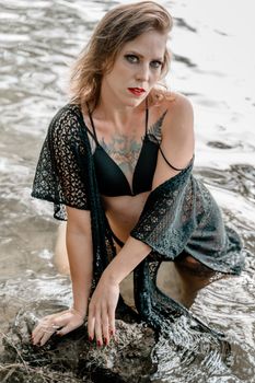 A woman in a black swimsuit and lace cover-up poses gracefully by a water's edge.
