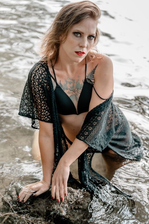 Free A woman in a black swimsuit and lace cover-up poses gracefully by a water's edge. Stock Photo