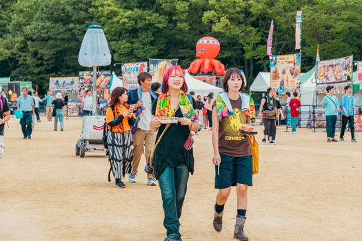 Colorful festival scene with people walking and enjoying street food under clear skies.