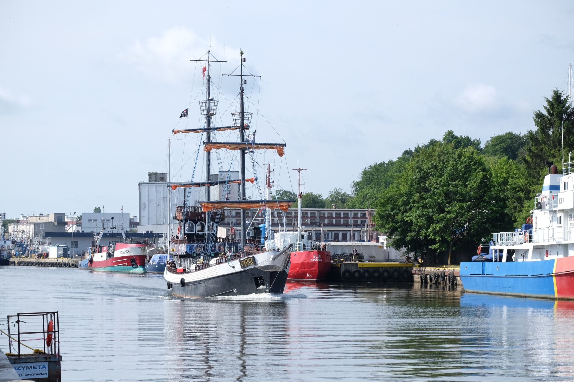 A historic sailing ship navigating a bustling, industrial harbor with diverse vessels.