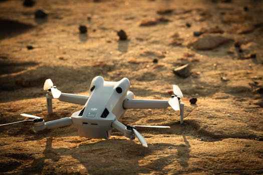 A drone rests on sandy ground, illuminated by the soft light of sunrise.