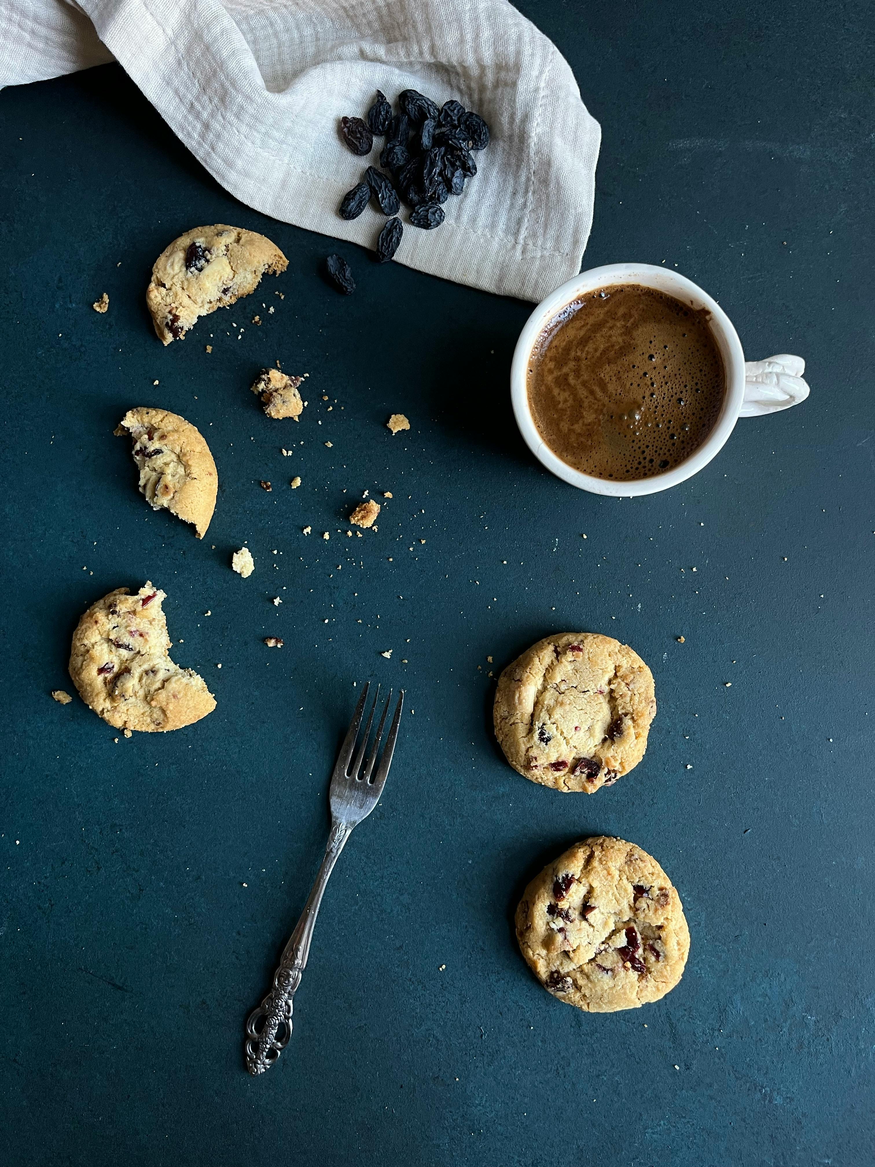 Flat lay of coffee and cookies on dark surface with raisins and fork.