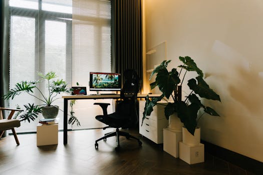 A stylish home office featuring large windows, a desk with a computer, and lush indoor plants.