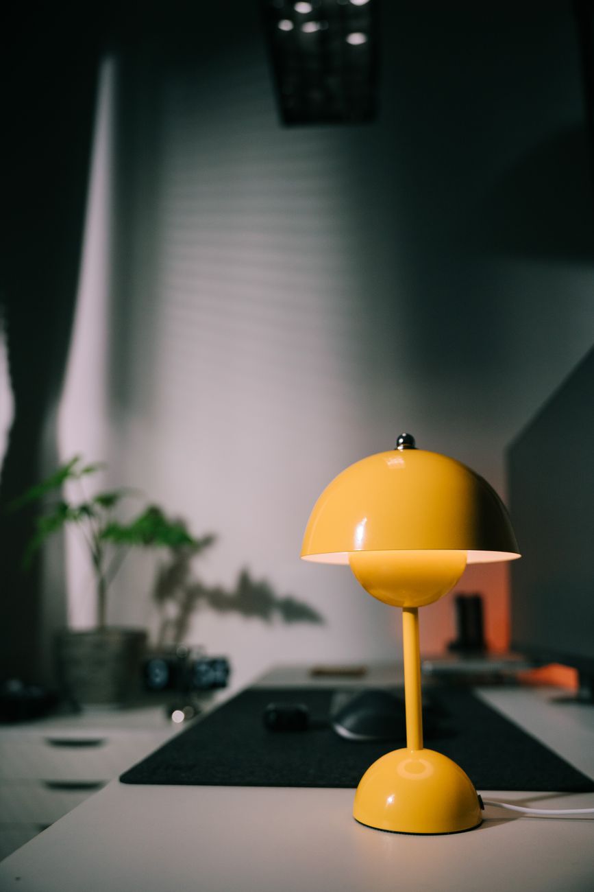 A bright yellow desk lamp illuminates a modern, minimalist workspace with a plant and shadows.