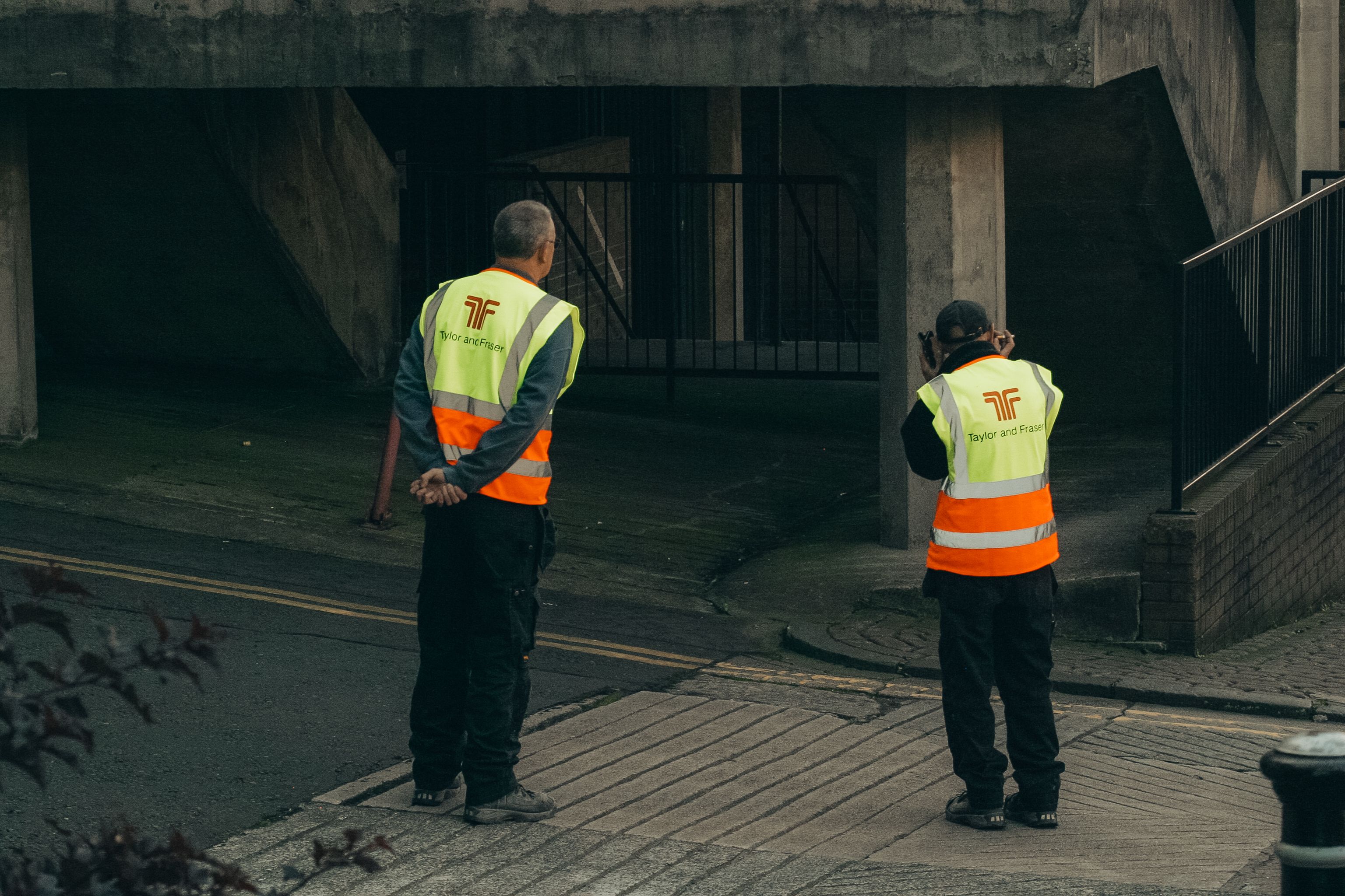 Two construction workers in reflective vests on a street beneath an overpass.