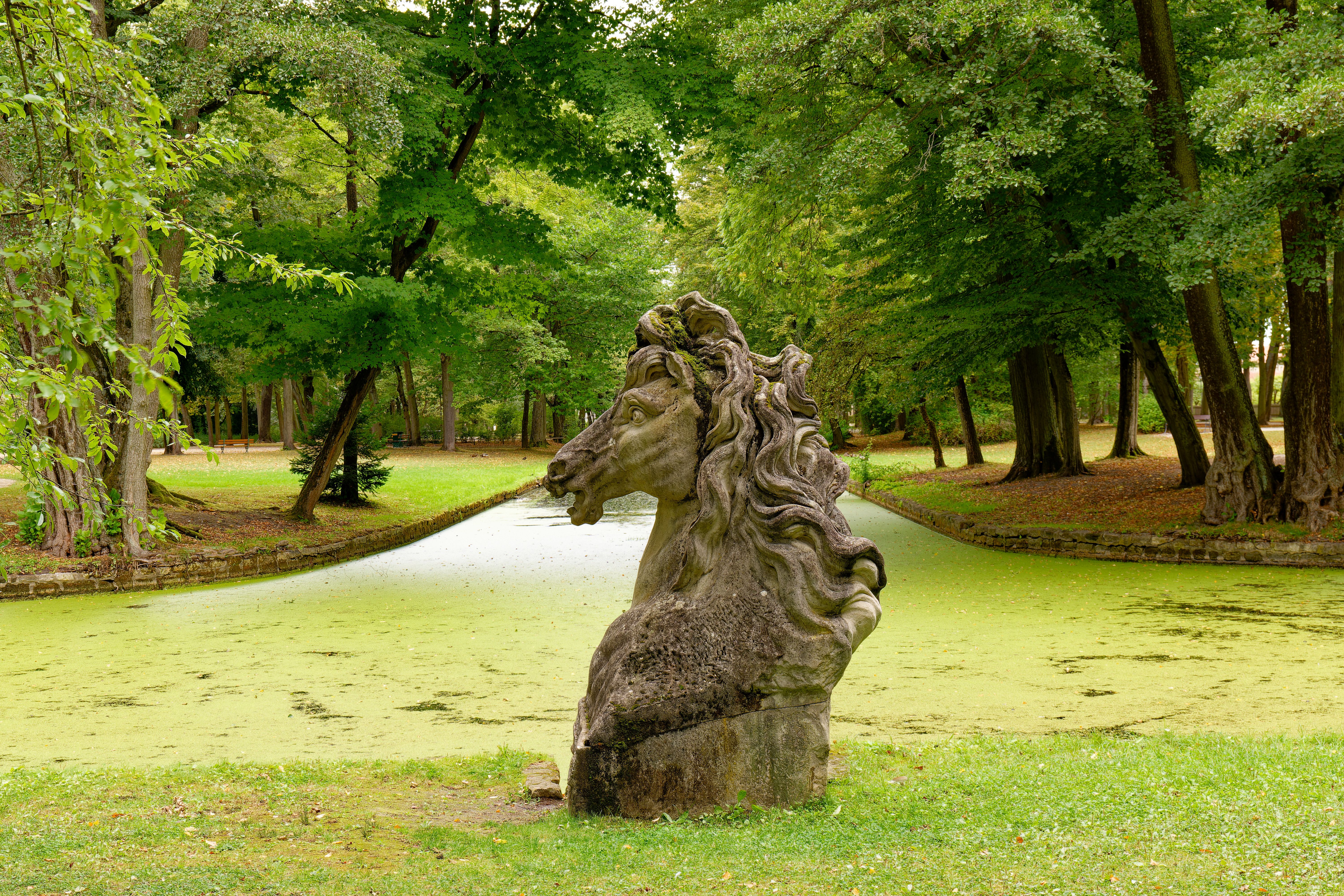A scenic view of a horsehead statue amidst vibrant green trees and a serene pond in Bayreuth, Germany.