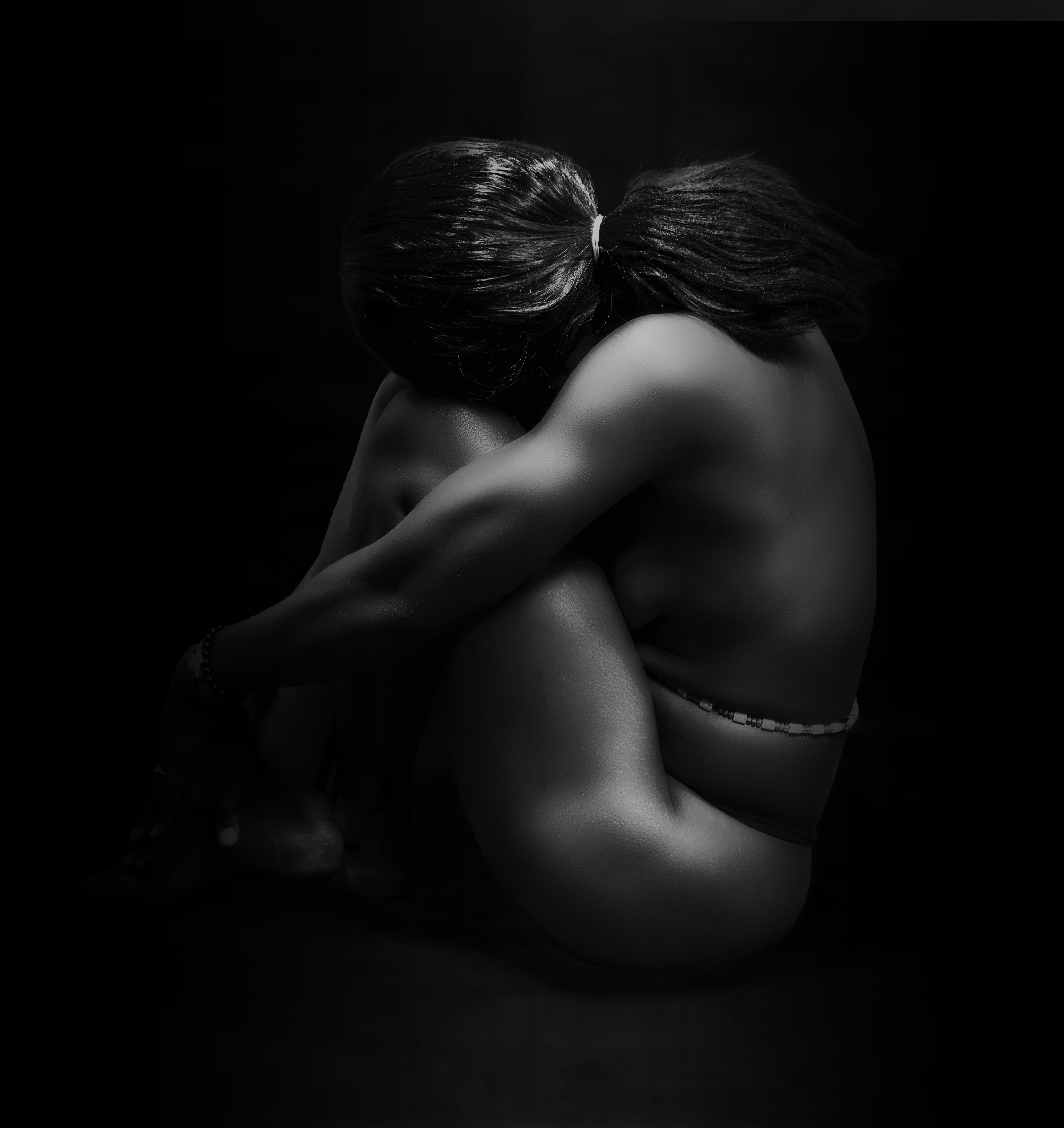 A dramatic black and white artistic nude portrait of a woman posed in a contemplative position, emphasizing curves and shadows.