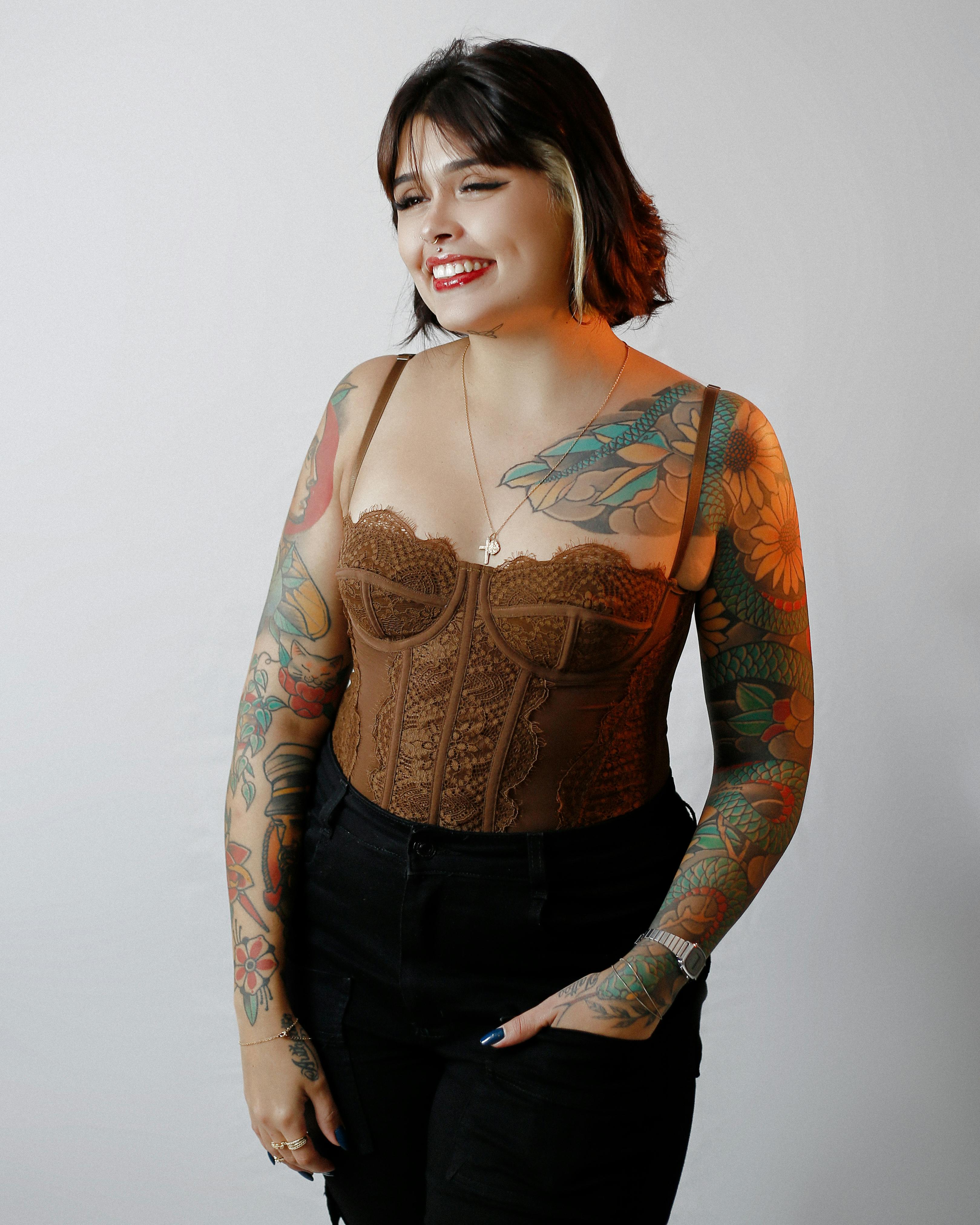 Confident woman with tattoos smiling, wearing a brown lace corset and black jeans.