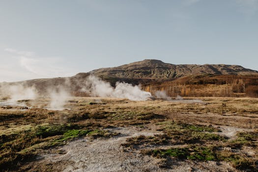 Captivating geothermal steam rising across a picturesque mountain landscape under clear skies.