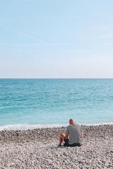 A solitary man sits on a pebble beach in Nice, France, gazing at the clear blue sea under a bright sky.