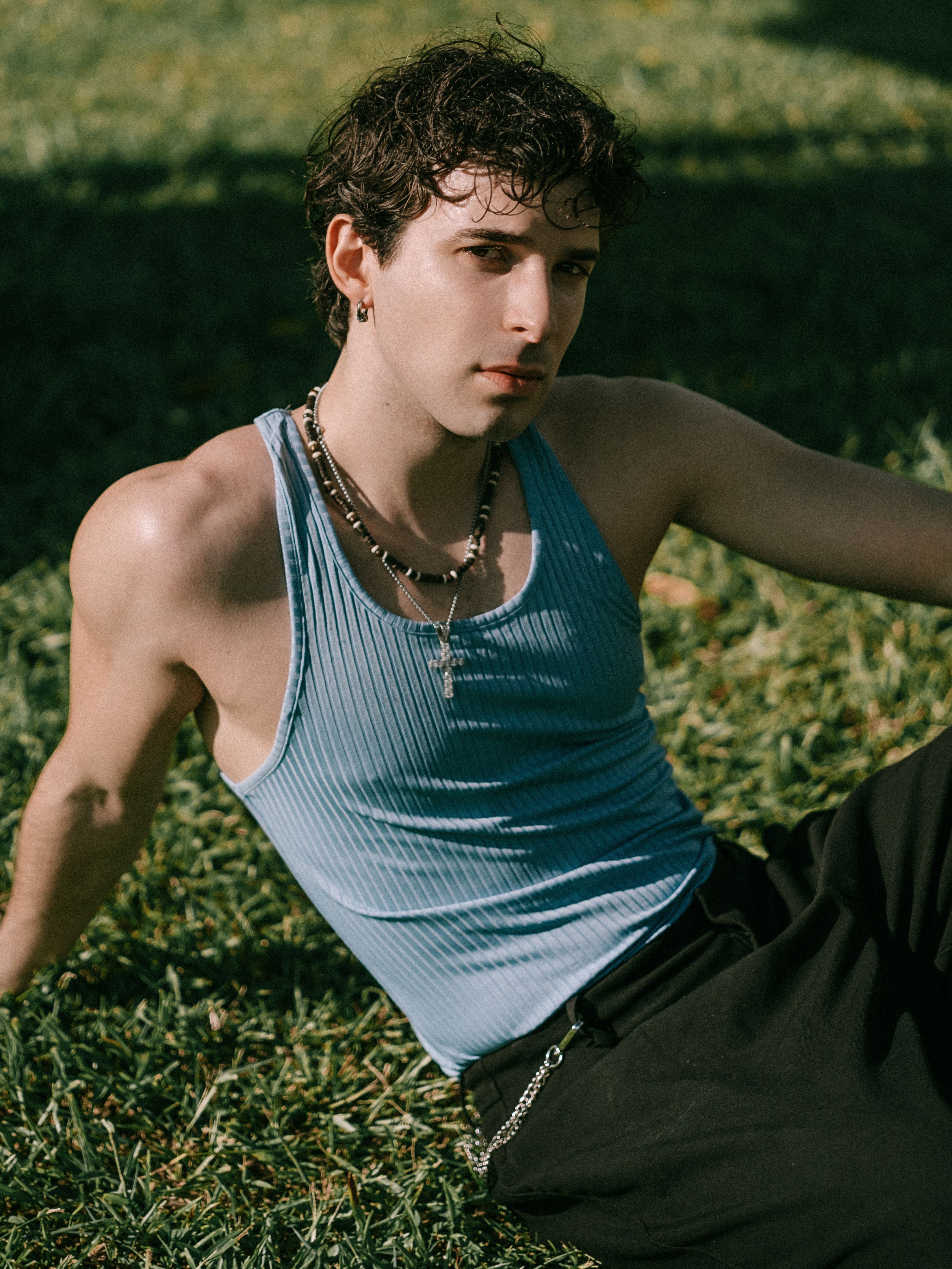 Young adult in blue tank top lounging on grass in daylight, wearing jewelry.