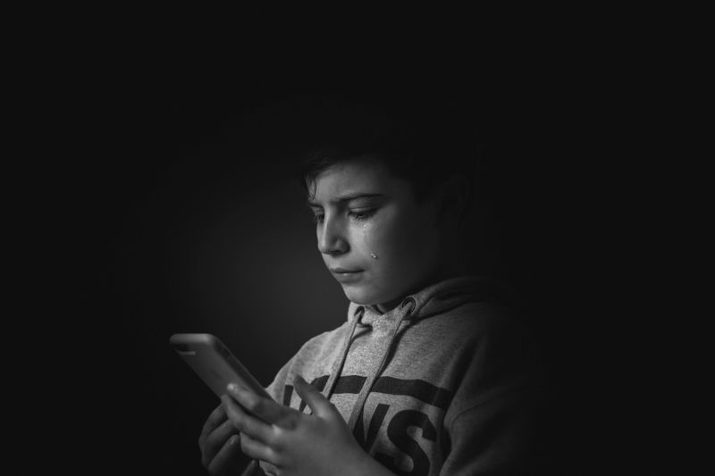 A monochromatic portrait of a boy holding a smartphone, expressing sadness