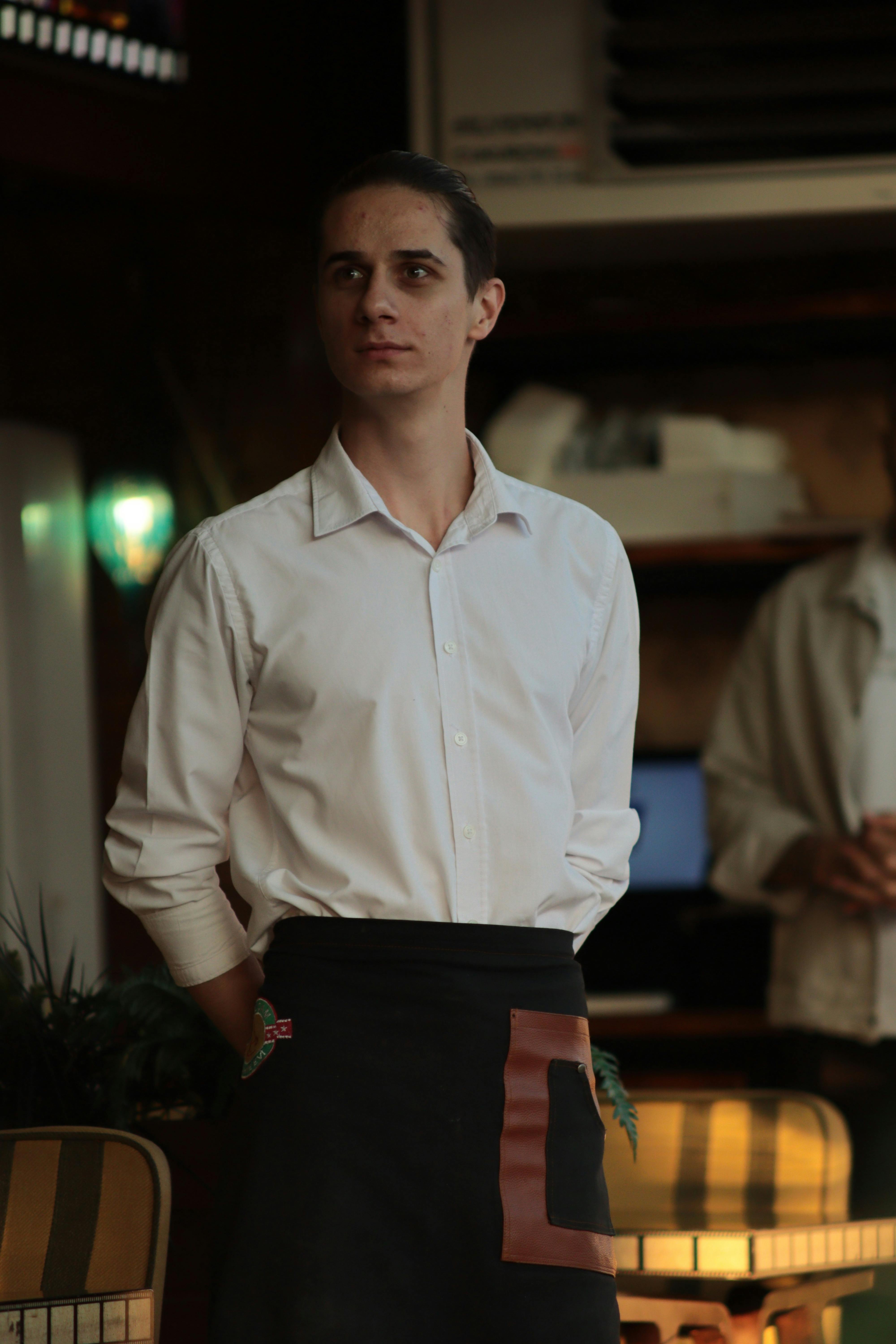 Young waiter in uniform standing indoors at a restaurant.