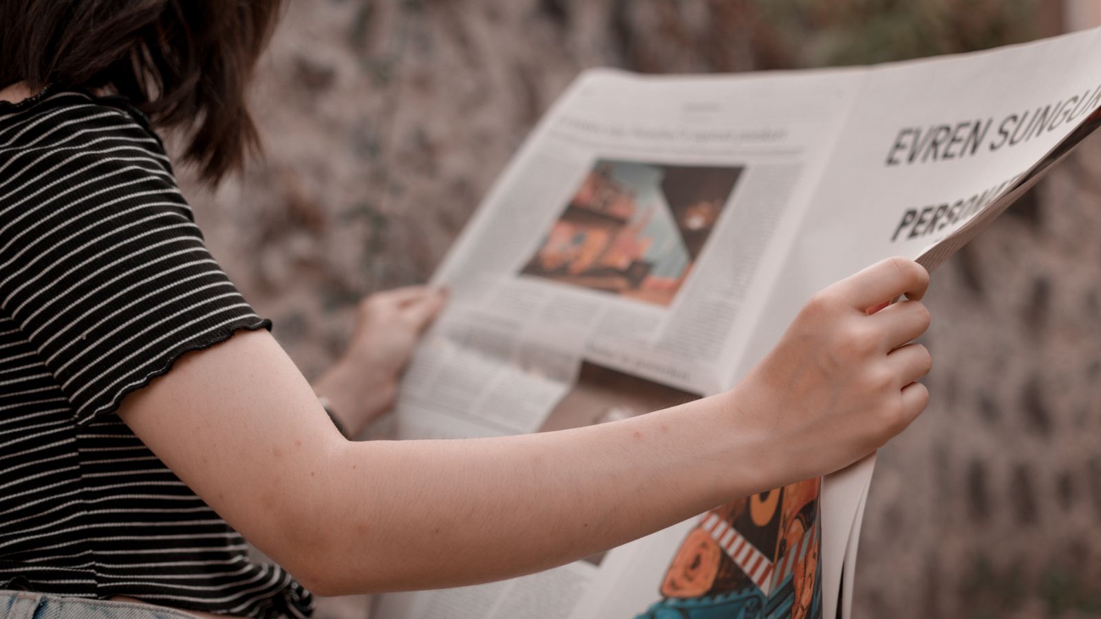 A woman holding a newspaper while sitting outside, enjoying a leisurely read.