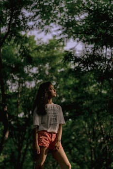 Woman in white shirt and shorts posing confidently in a sunlit forest.