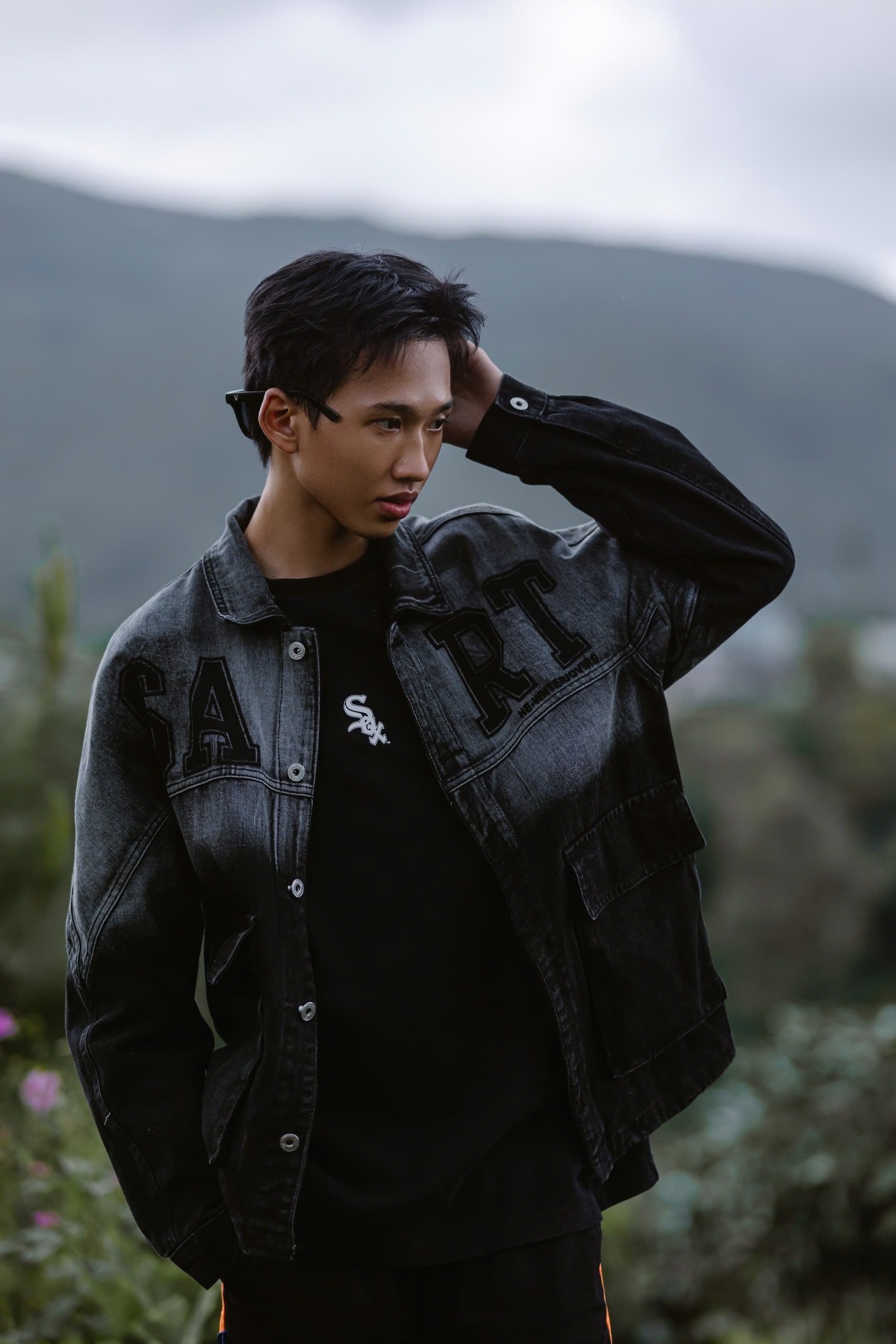 Young man posing outdoors in a stylish denim jacket with moody mountain backdrop.