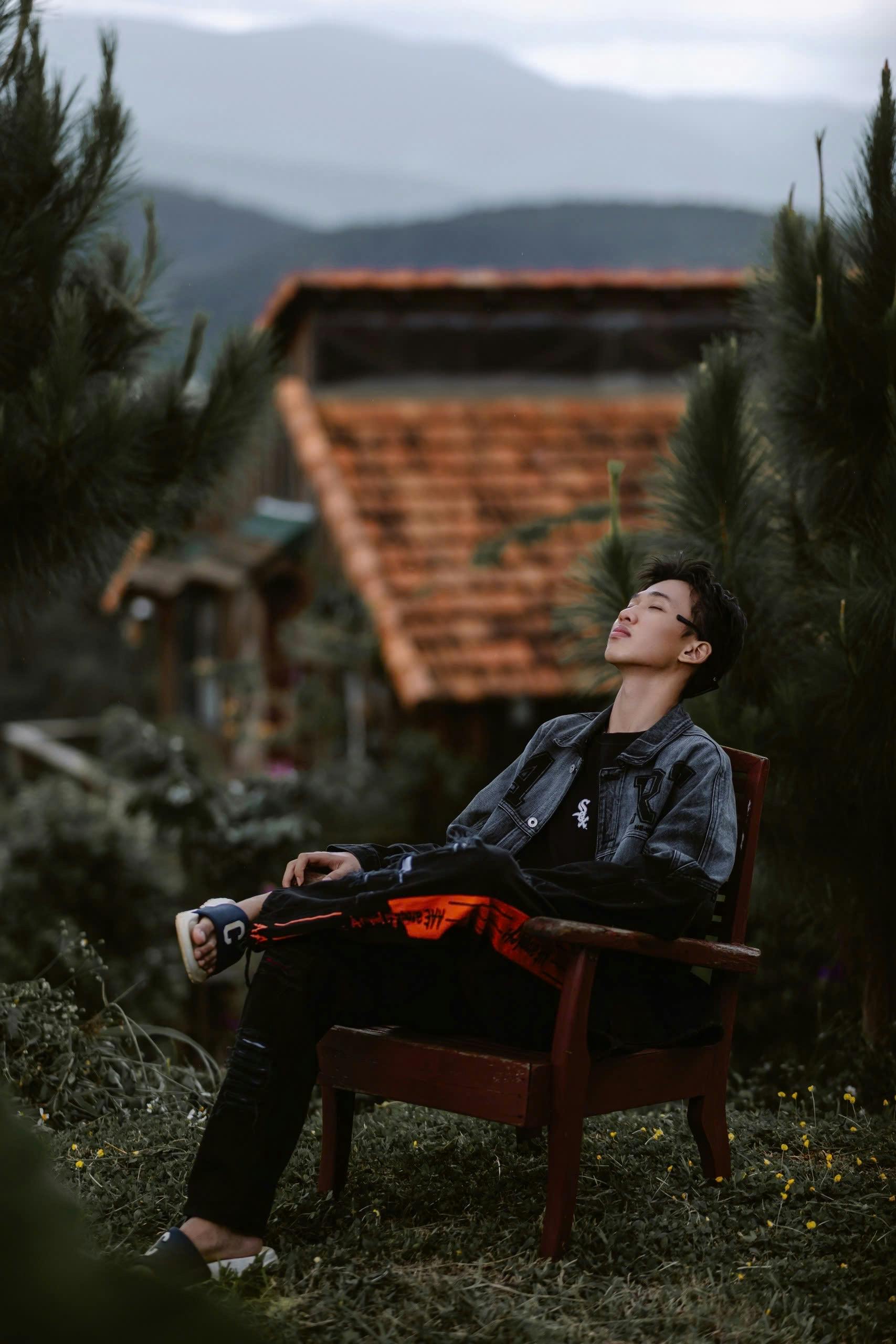 Man relaxing in a chair amidst lush greenery with mountain views and a rustic house.