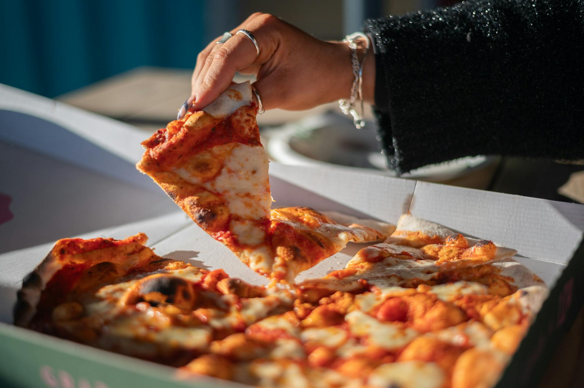 A close-up of a person's hand taking a slice of cheese pizza from a box outdoors.