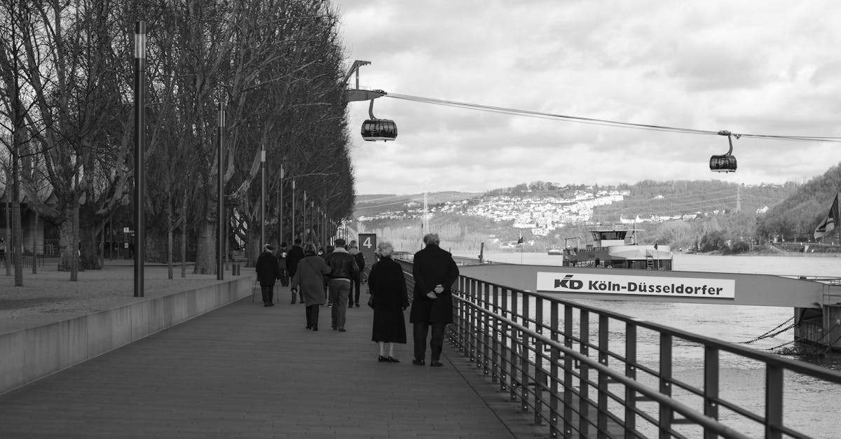 Black and white view of people strolling along the Koblenz riverfront with cable cars overhead.