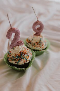 Two cupcakes with colorful sprinkles and candles depicting the number 29.